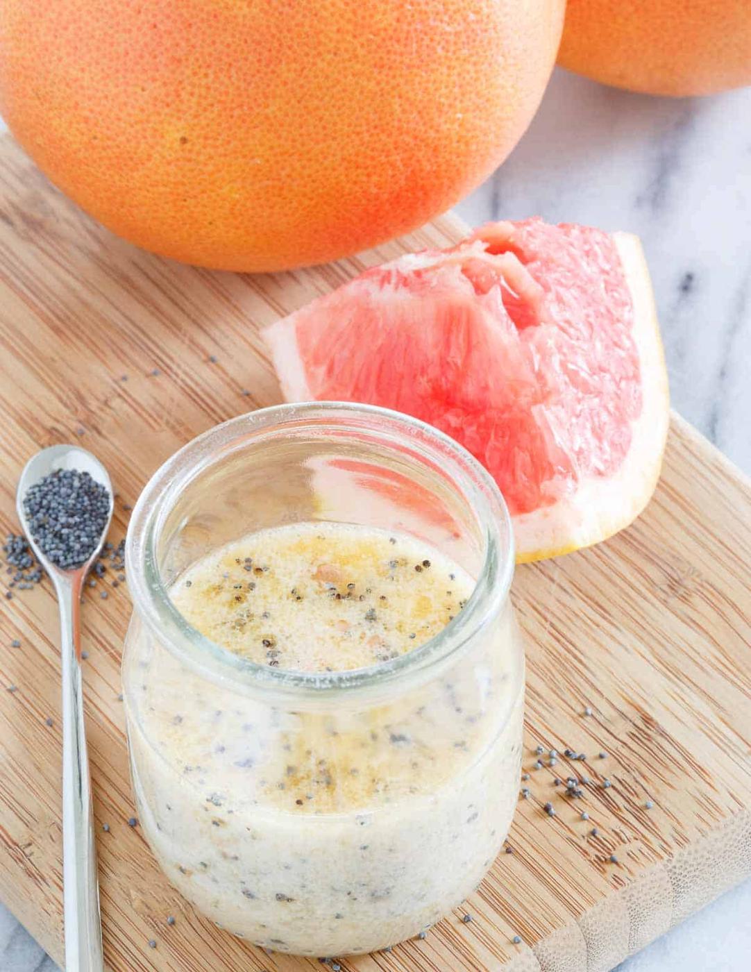 A small glass jar with creamy citrus dressing containing poppy seeds sits on a wooden board next to a spoon of poppy seeds and a wedge of pink grapefruit. Whole grapefruits are in the background.