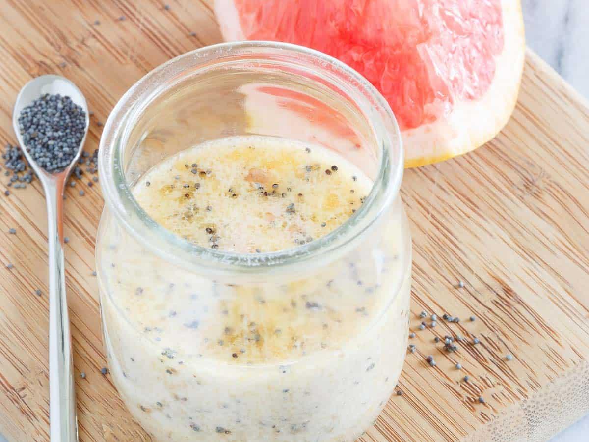 A glass jar of creamy citrus poppy seed dressing sits on a wooden board next to a sliced grapefruit and a spoonful of poppy seeds, with some seeds scattered on the board.