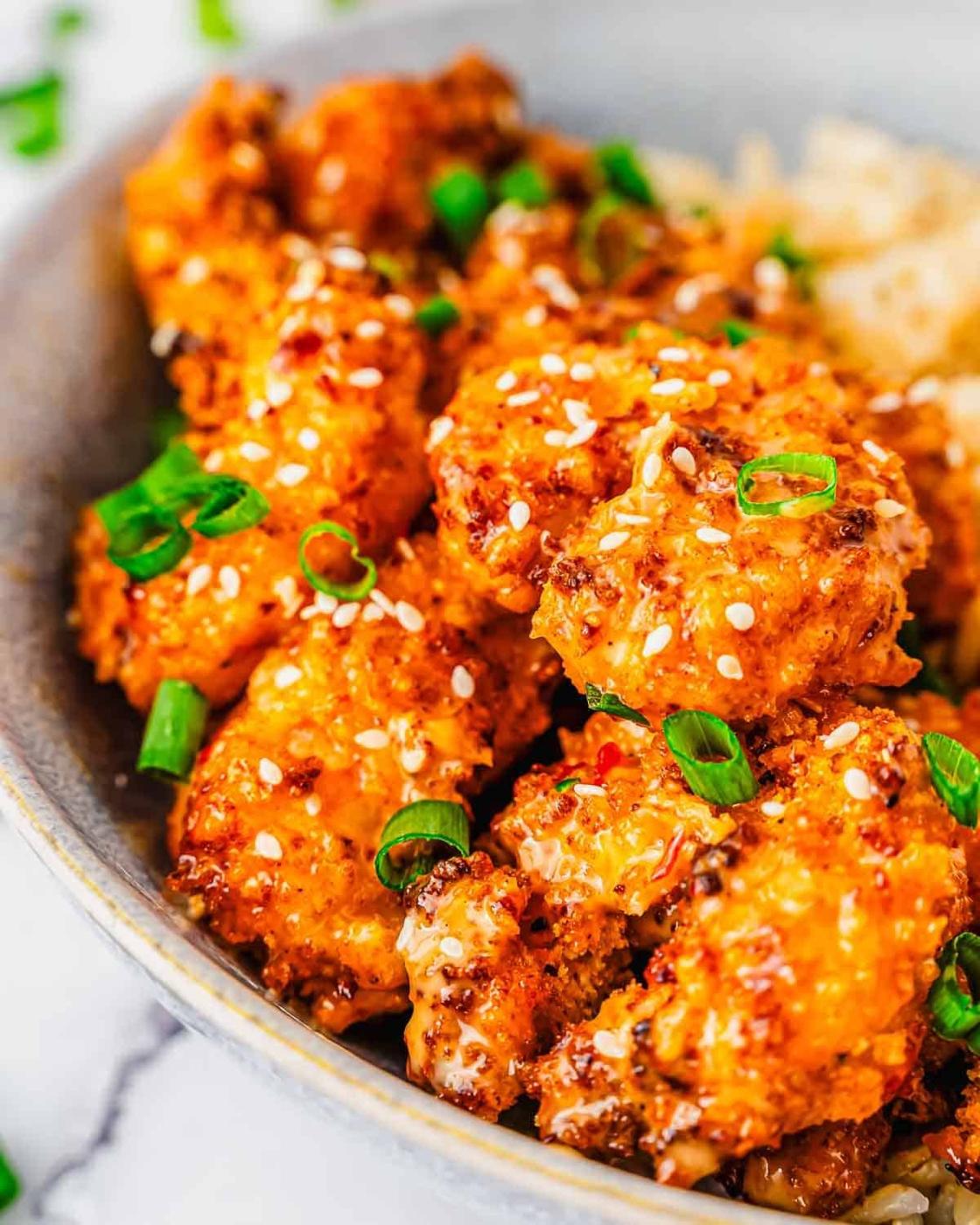 A bowl of crispy, golden-brown air fryer bang bang shrimp pieces topped with sesame seeds and chopped green onions, served over a bed of rice.
