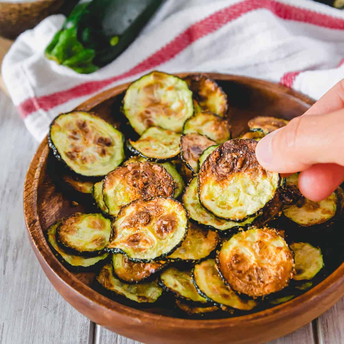 Garlic zucchini chips made in the air fryer on a serving plate with someone grabbing a chip to eat.