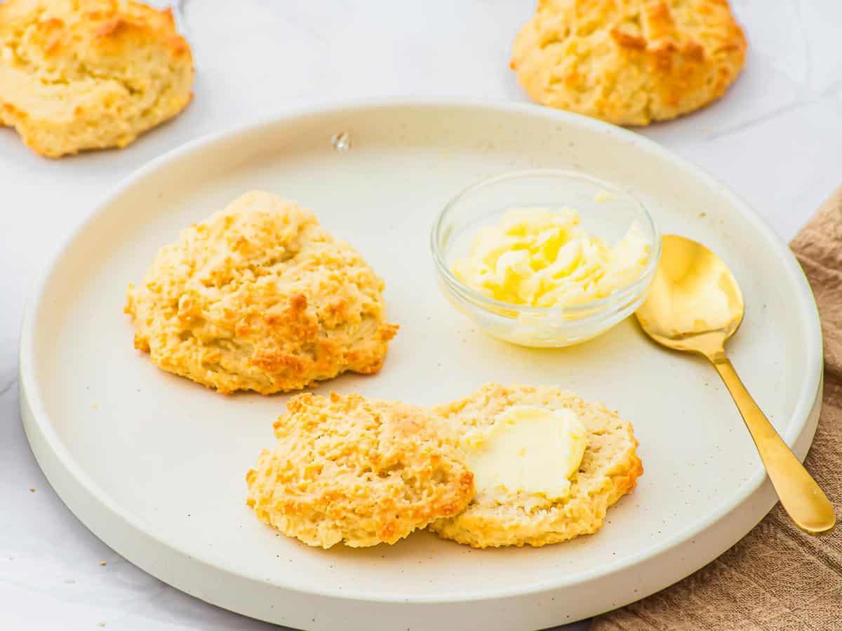 A round plate with two golden-brown almond flour biscuits, one split and spread with butter, next to a small glass bowl of butter and a gold spoon. More biscuits are visible in the background.