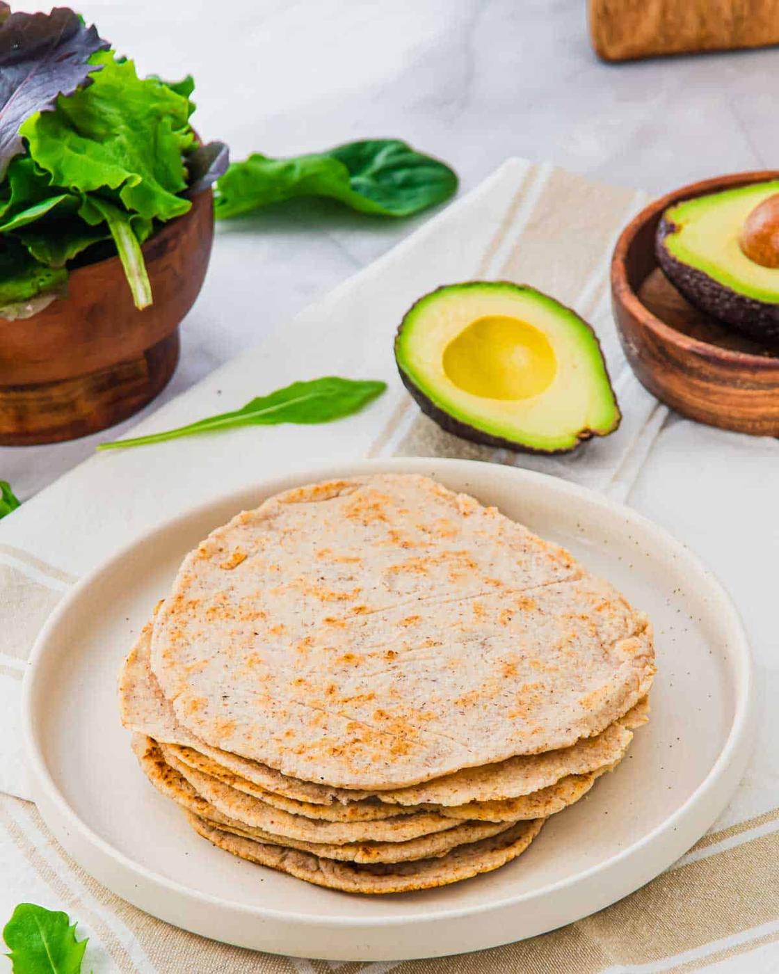 A stack of almond flour tortillas on a white plate, with a halved avocado, a bowl of mixed greens, and spinach leaves on a light-colored surface in the background.