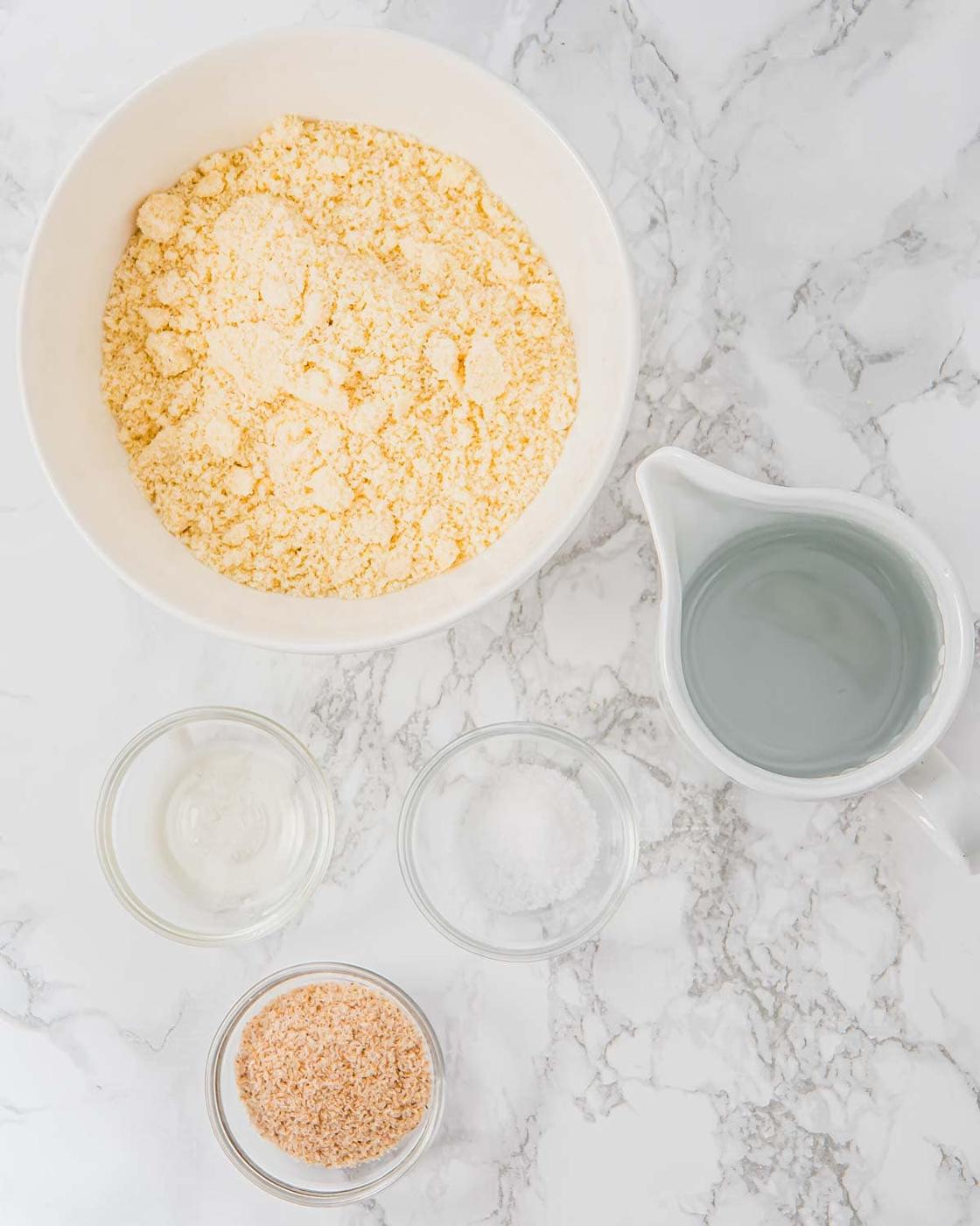 A bowl of almond flour, a pitcher of water, and three small bowls with vinegar, salt, and psyllium husk are arranged on a white marble surface.
