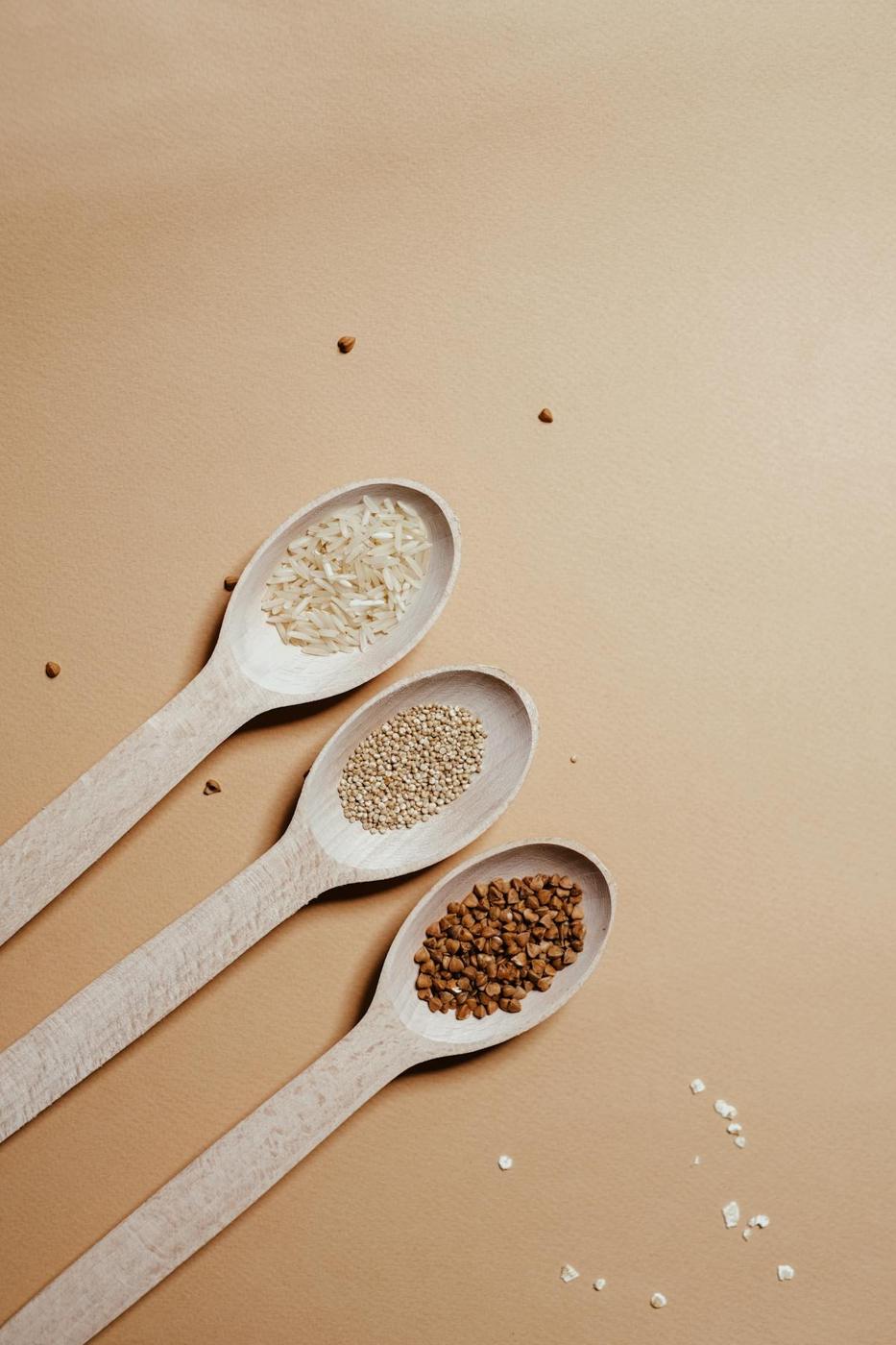 Three wooden spoons containing ancient grains, sesame seeds, and cloves on a beige surface.