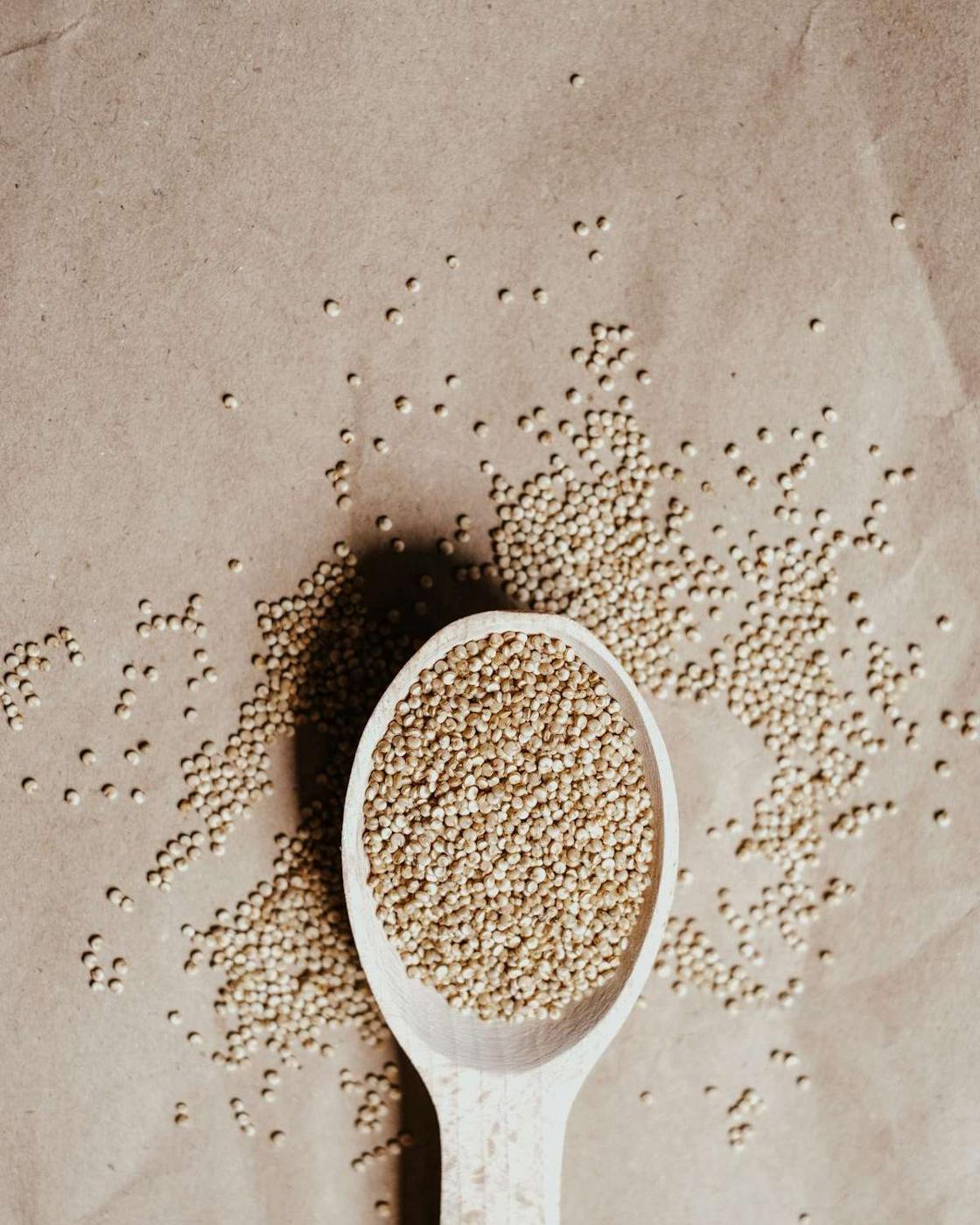 A wooden spoon overflowing with ancient grains, specifically quinoa seeds, on a brown paper surface.