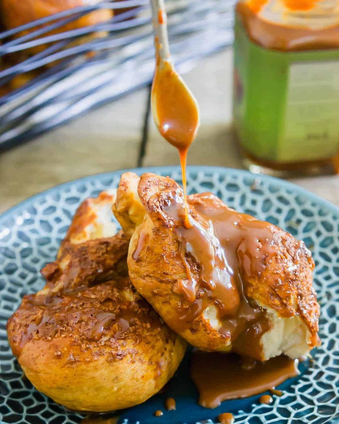 A spoon drizzles caramel sauce over two golden brown pretzel twists on a blue patterned plate. A jar of caramel sauce and a wire rack appear blurred in the background.