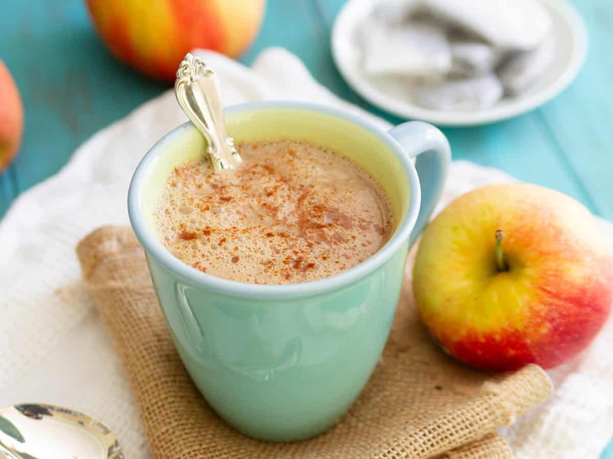 A light blue mug filled with a tea latte drink, topped with cinnamon, is placed on a napkin next to a red and yellow apple. A spoon rests inside the mug, and a plate of teabags is visible in the background.
