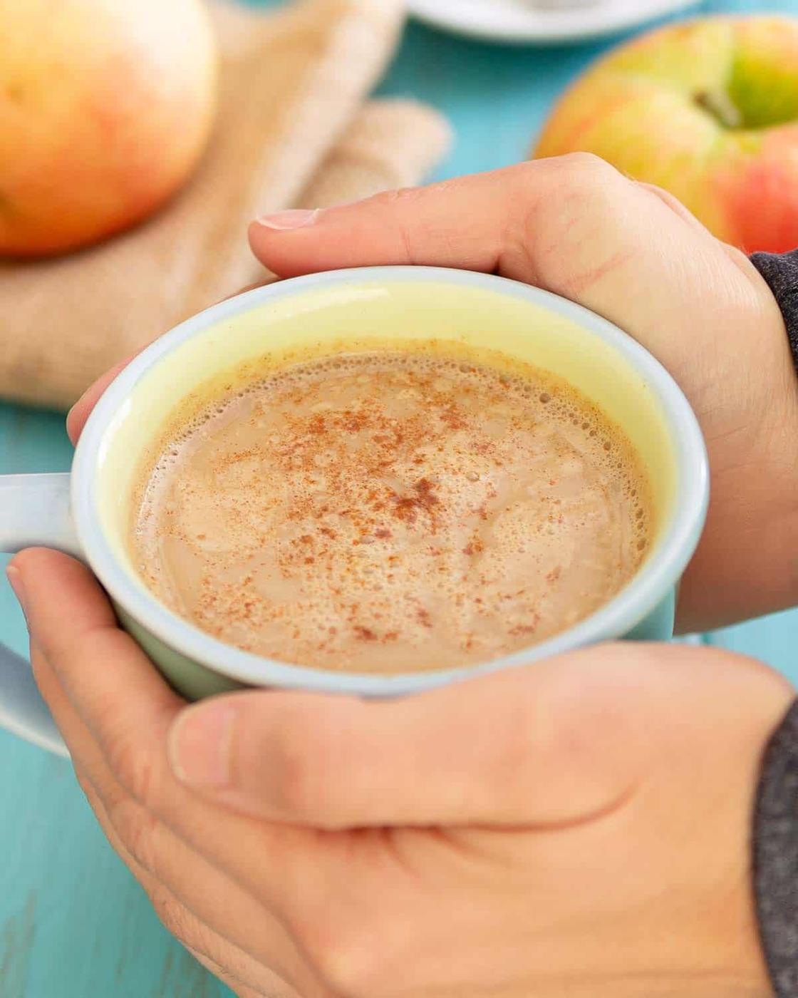 Hands holding a blue mug filled with a warm tea latte, topped with cinnamon. An apple and a cloth are in the background.