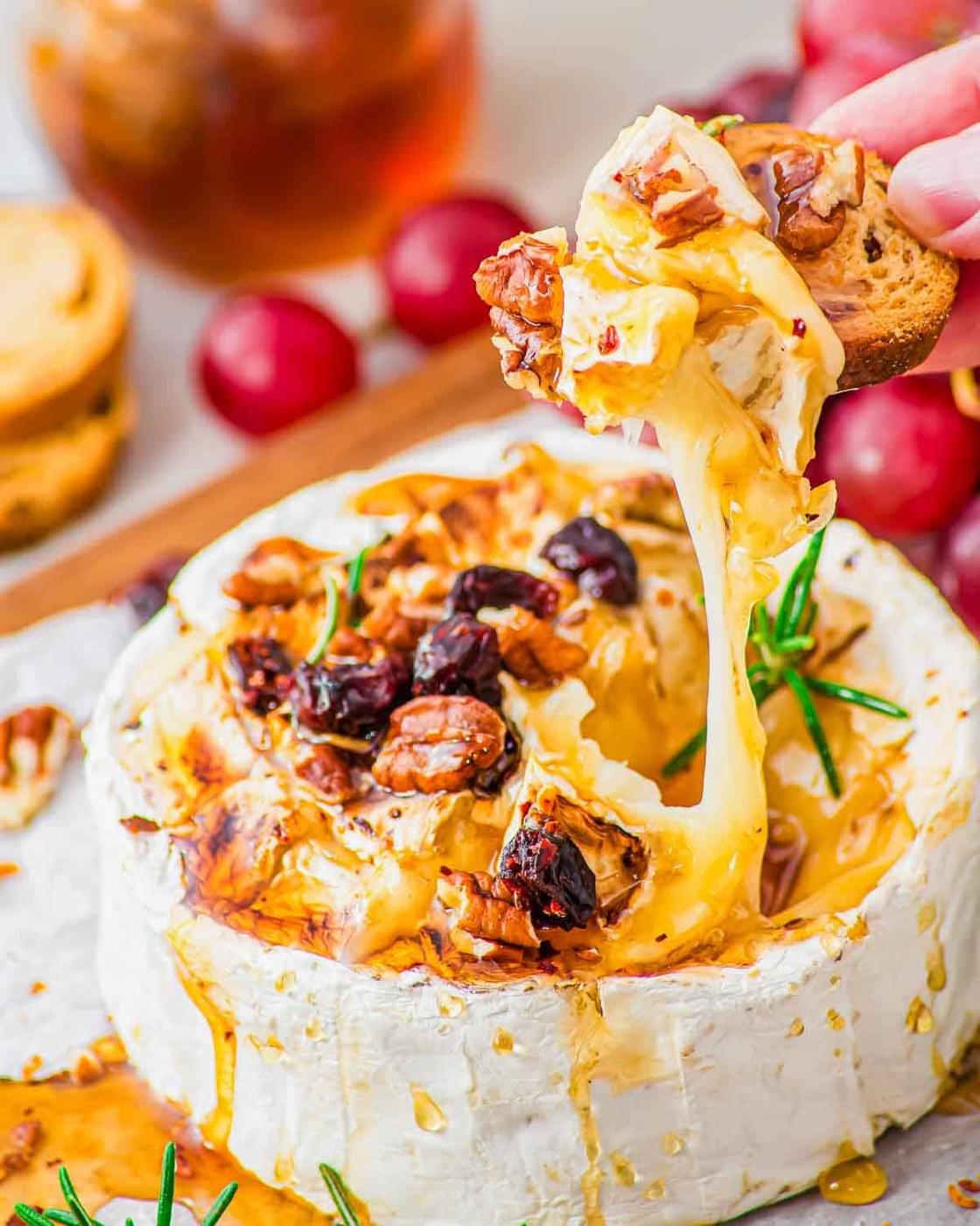 A hand dips a toasted baguette slice into warm, gooey baked brie topped with honey, nuts, and dried cranberries. Fresh rosemary and grapes are seen nearby on the serving board.