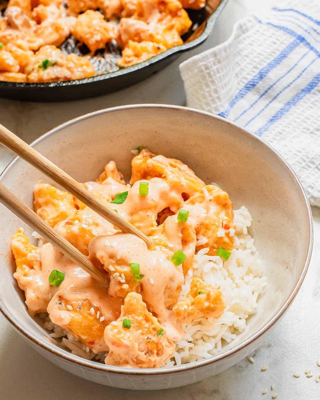 A bowl of white rice topped with crispy pan-fried cauliflower pieces garnished with bang bang sauce and chopped green onions. A pair of chopsticks is lifting a piece of chicken. A skillet with more cauliflower is in the background, next to a striped cloth.
