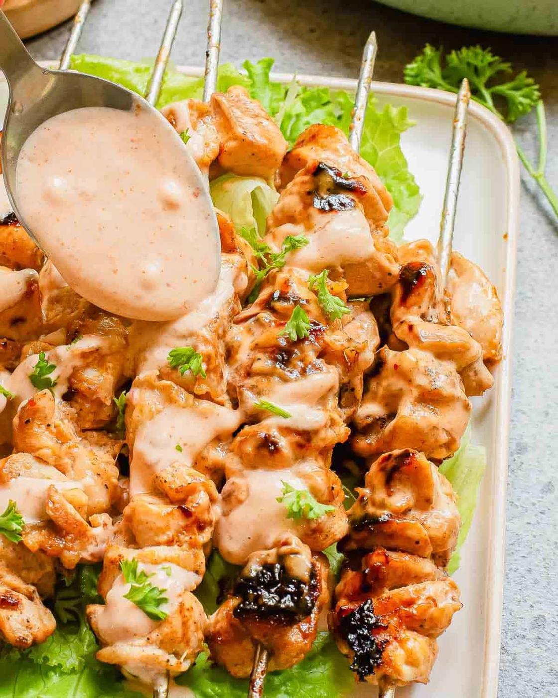 Spooning extra sauce on top of chicken skewers on a platter with lettuce.