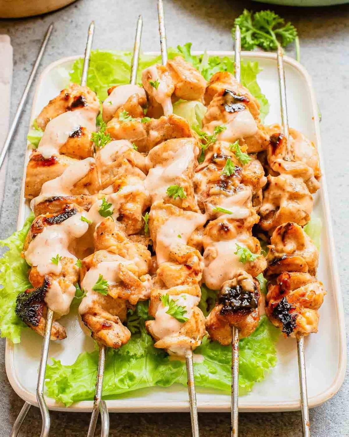Chicken skewers lined up on a serving plate drizzled with creamy bang bang sauce.