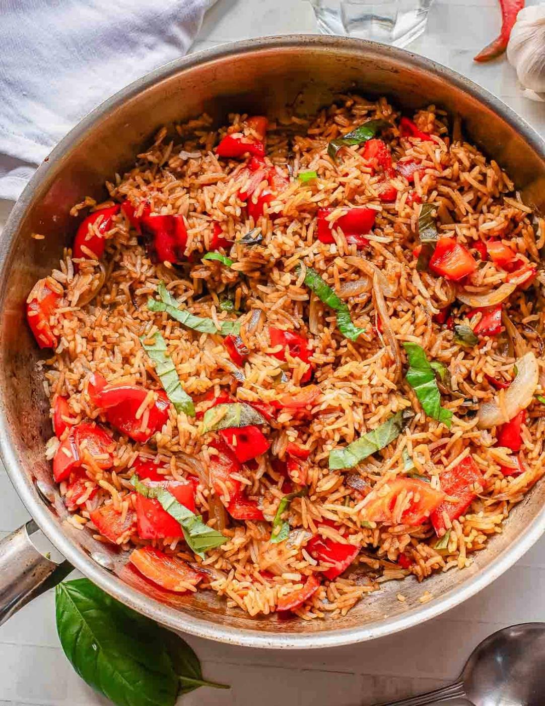 A skillet filled with basil fried rice, chopped tomatoes, sliced onions, and fresh basil strips, all mixed together and seasoned, sits on a light-colored surface with a basil leaf nearby.