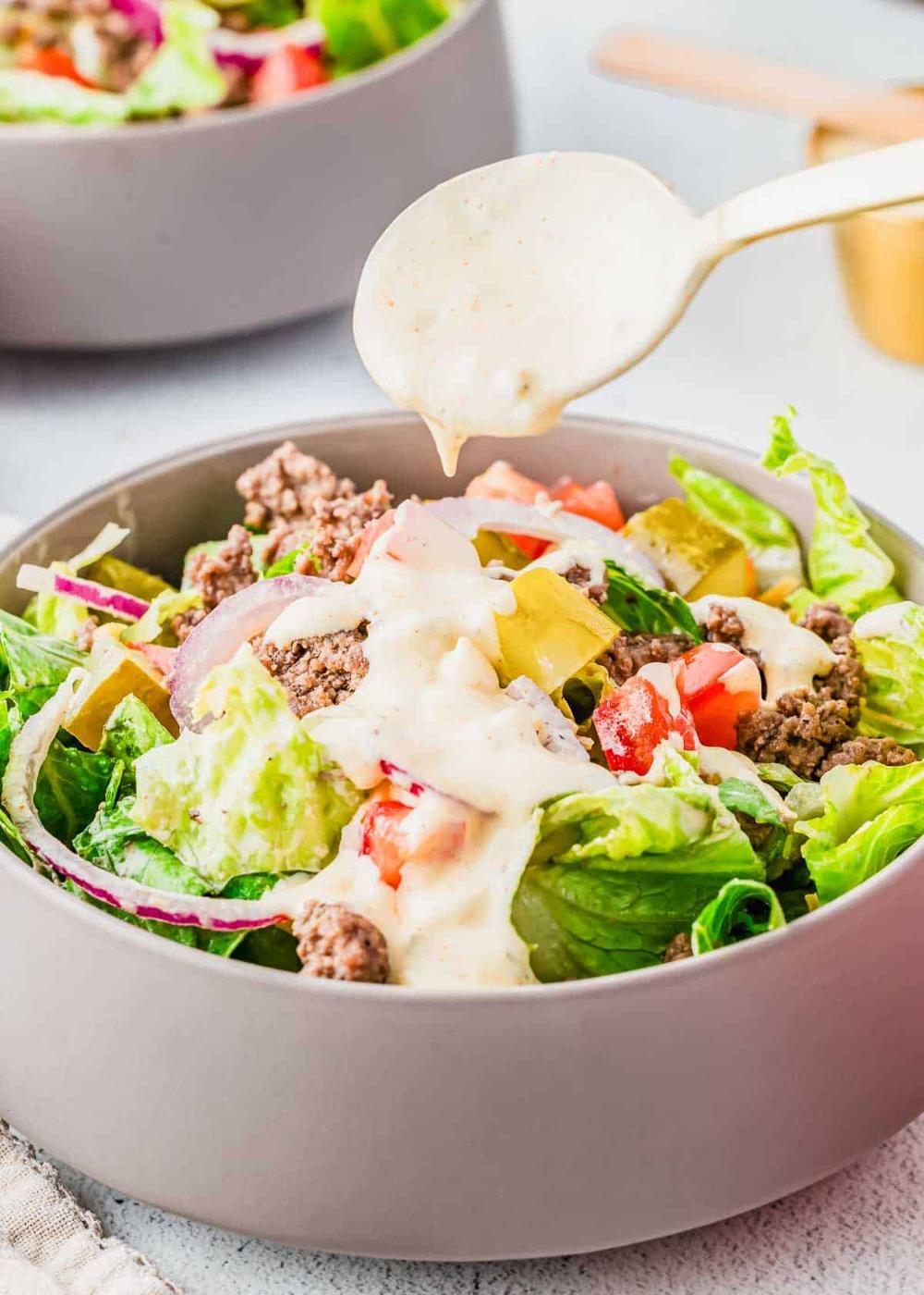 A gray bowl filled with a salad containing ground beef, lettuce, tomato, pickles, and red onions, with creamy dressing being drizzled on top from a spoon above.