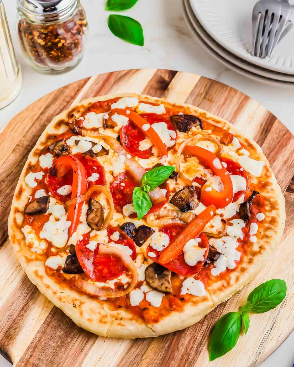 A freshly baked pizza with pepperoni, onions, bell peppers, mushrooms, and melted cheese sits on a wooden cutting board next to a jar of seasoning and a stack of white plates with utensils.