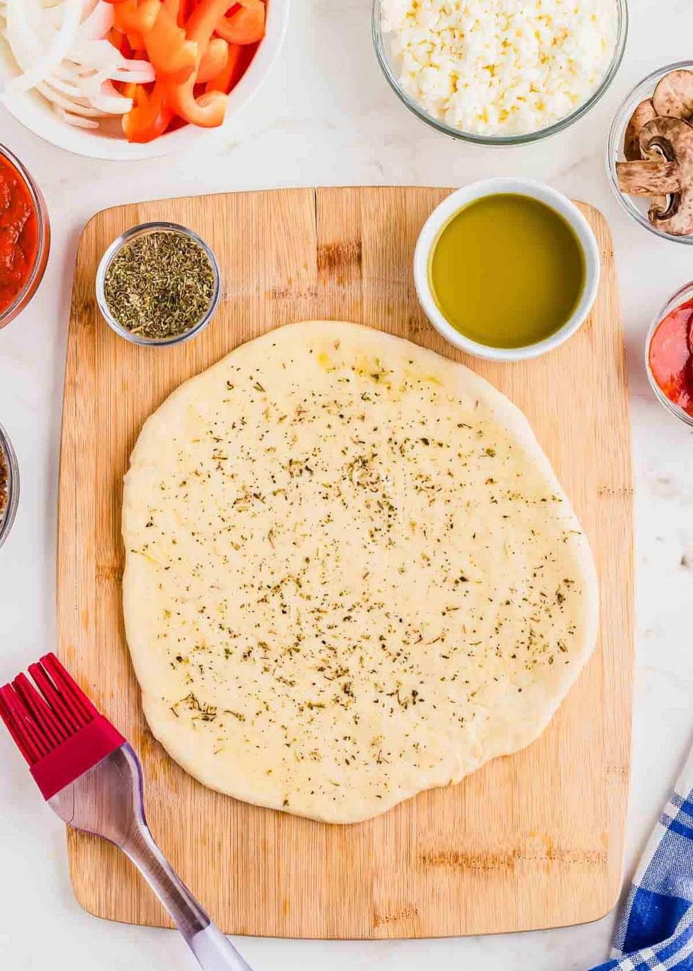 Flat pizza dough with herbs on a wooden board, surrounded by bowls containing olive oil, spices, vegetables, mushrooms, cheese, and a pastry brush on the side.
