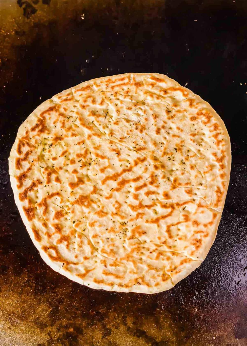 Cooked pizza dough with a golden-brown, slightly charred surface is shown on a Blackstone griddle surface. The crust appears to have a crispy texture with light seasonings sprinkled on top.
