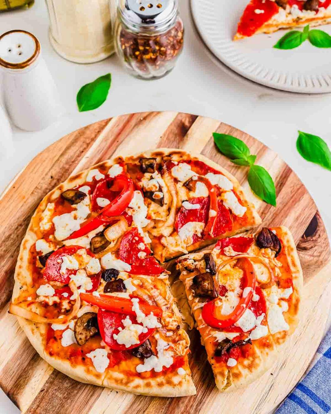 A pizza cooked on the Blackstone griddle topped with mushrooms, red bell peppers, pepperoni, and cheese sits on a wooden board. A slice has been removed. Basil leaves and seasoning containers are visible around it.