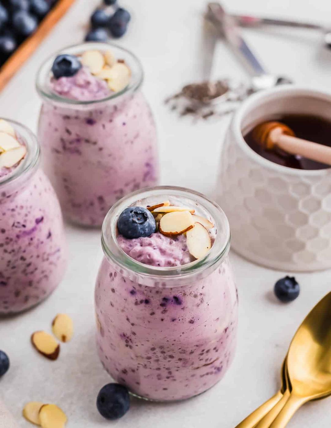 Three small glass jars filled with blueberry chia pudding, topped with almond slices and blueberries, are placed in the foreground. A honey jar, spoon, and other ingredients are in the background.