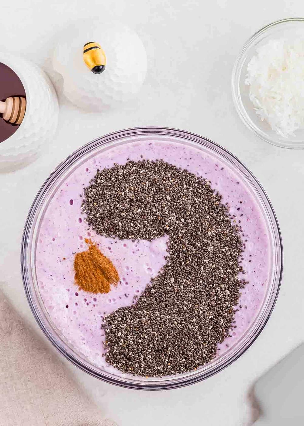 A glass bowl filled with a blended blueberry mixture topped with a large amount of chia seeds arranged in a swirling shape, a small pile of cinnamon, with honey and shredded coconut nearby.