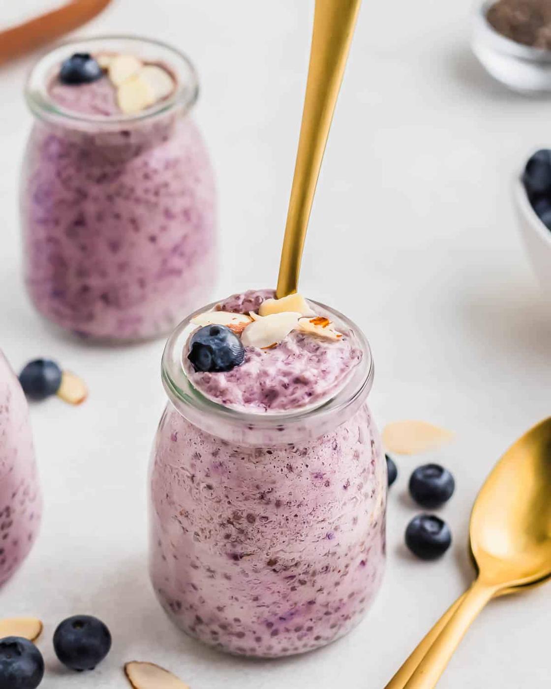Three jars of blueberry chia pudding topped with fresh blueberries and almond slices, with a golden spoon inserted in one. Scattered blueberries and almond slices are on the surface.