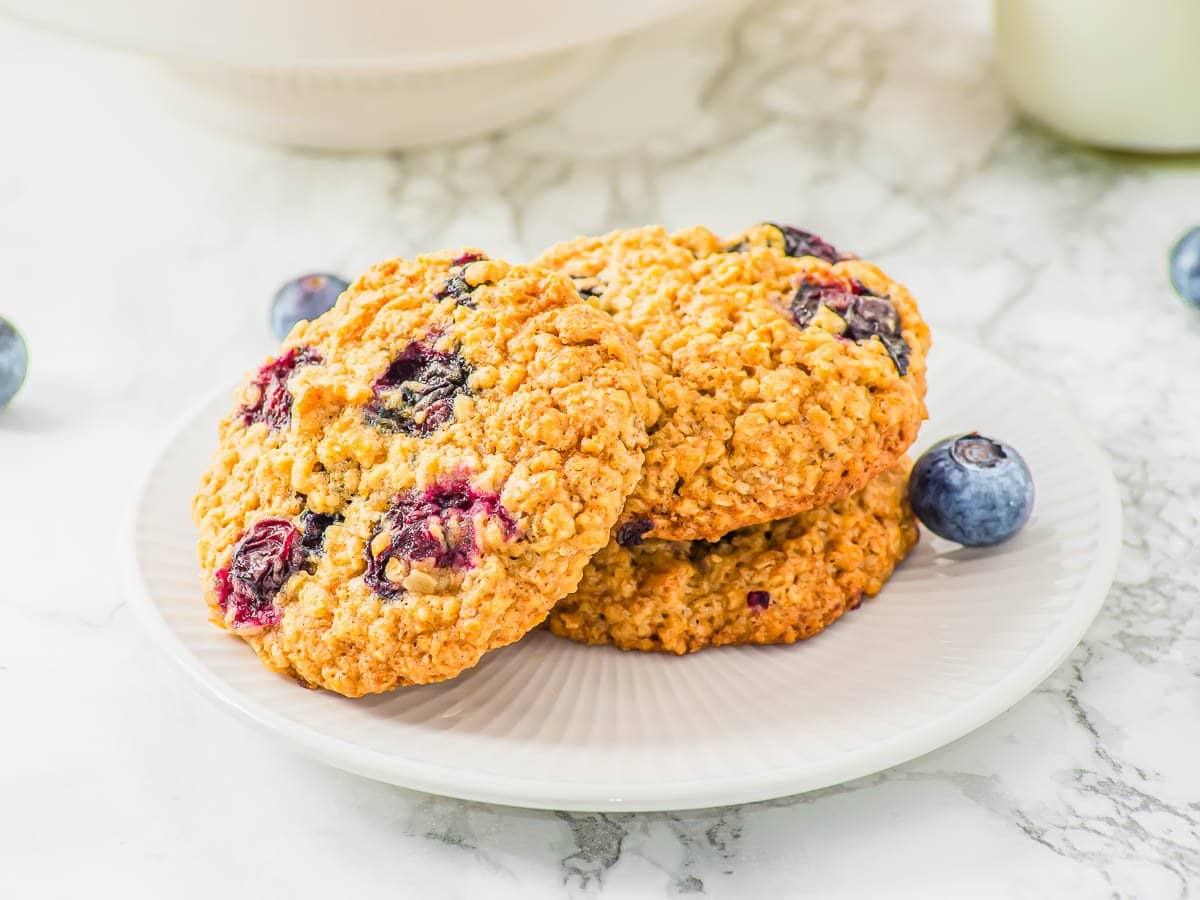 Three oatmeal blueberry cookies on a white plate, set on a marble countertop. Blueberries are scattered around the plate, adding a fresh touch to the scene.