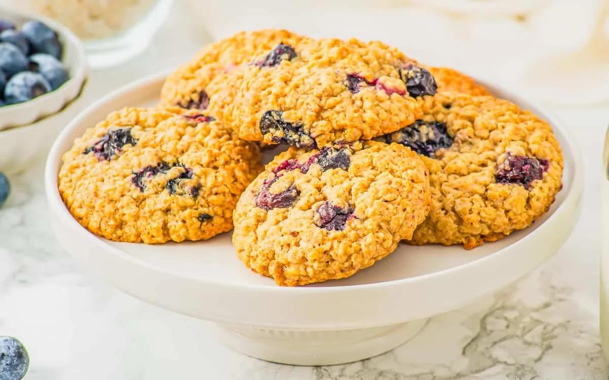 A white plate displays a stack of blueberry oatmeal cookies. In the background, there are fresh blueberries in a bowl, all set on a marble surface. The scene is brightly lit, highlighting the cookies' texture.