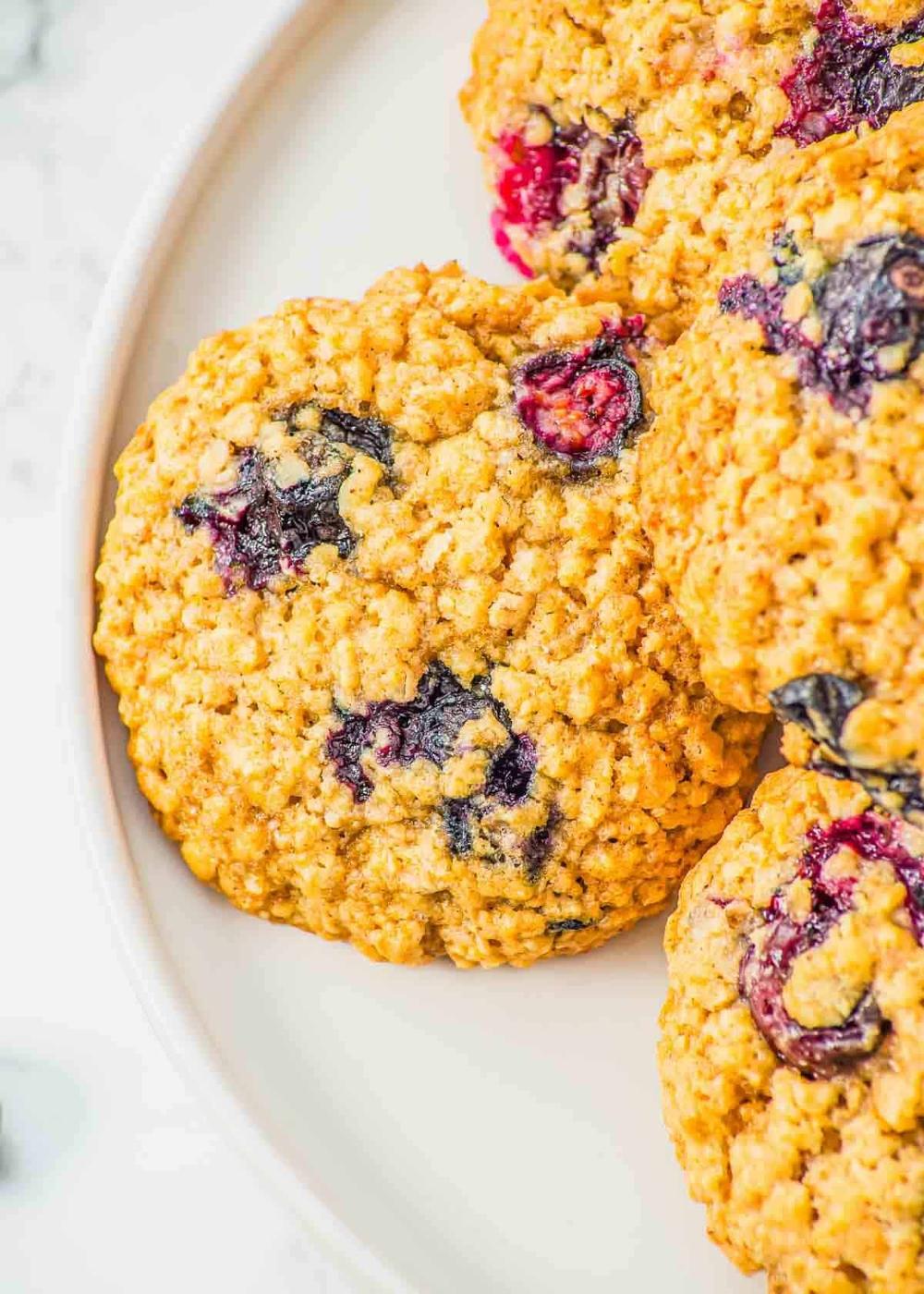 Close-up of three oatmeal cookies with fresh blueberries on a white plate. The cookies have a golden-brown, textured surface with colorful berries embedded. The background is a marble surface.