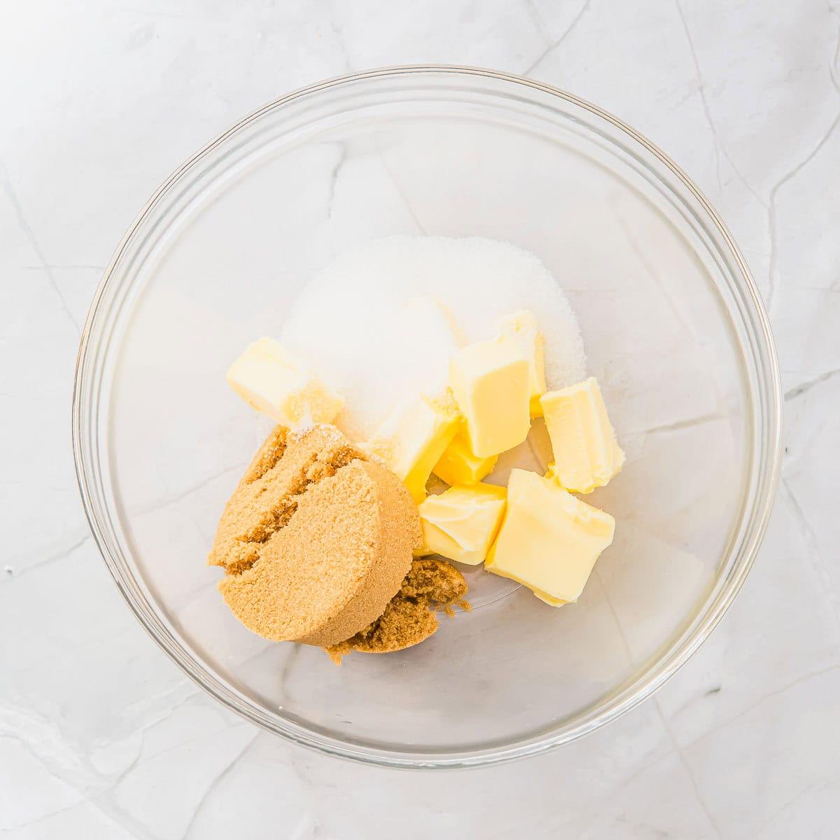 A glass bowl containing cubes of butter, a mound of brown sugar, and white granulated sugar, placed on a light-colored marble surface.