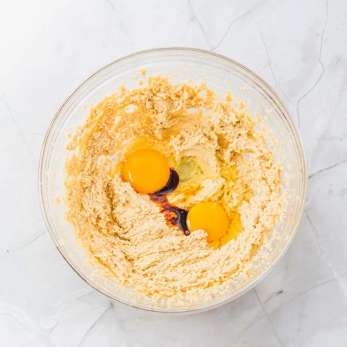 A glass bowl on a marble surface contains beaten sugar and butter mixture, with two raw eggs and vanilla extract in the center. The ingredients are ready to be mixed for baking.