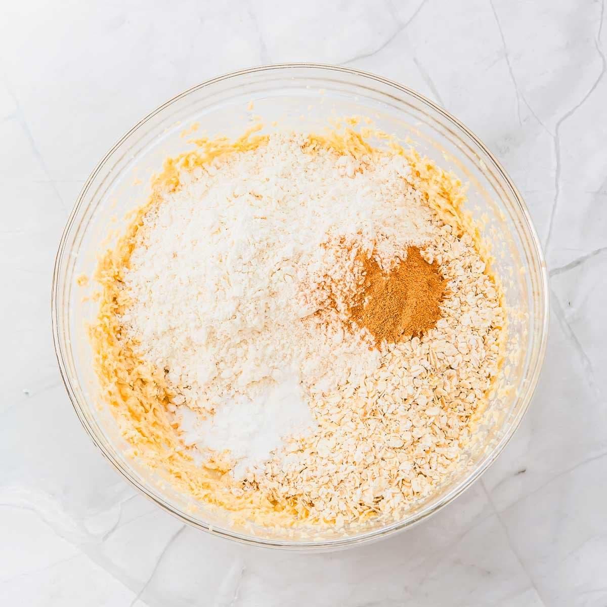 A glass bowl on a marble surface filled with ingredients for baking cookies, including flour, oats, baking soda, and cinnamon on top of a creamy mixture.