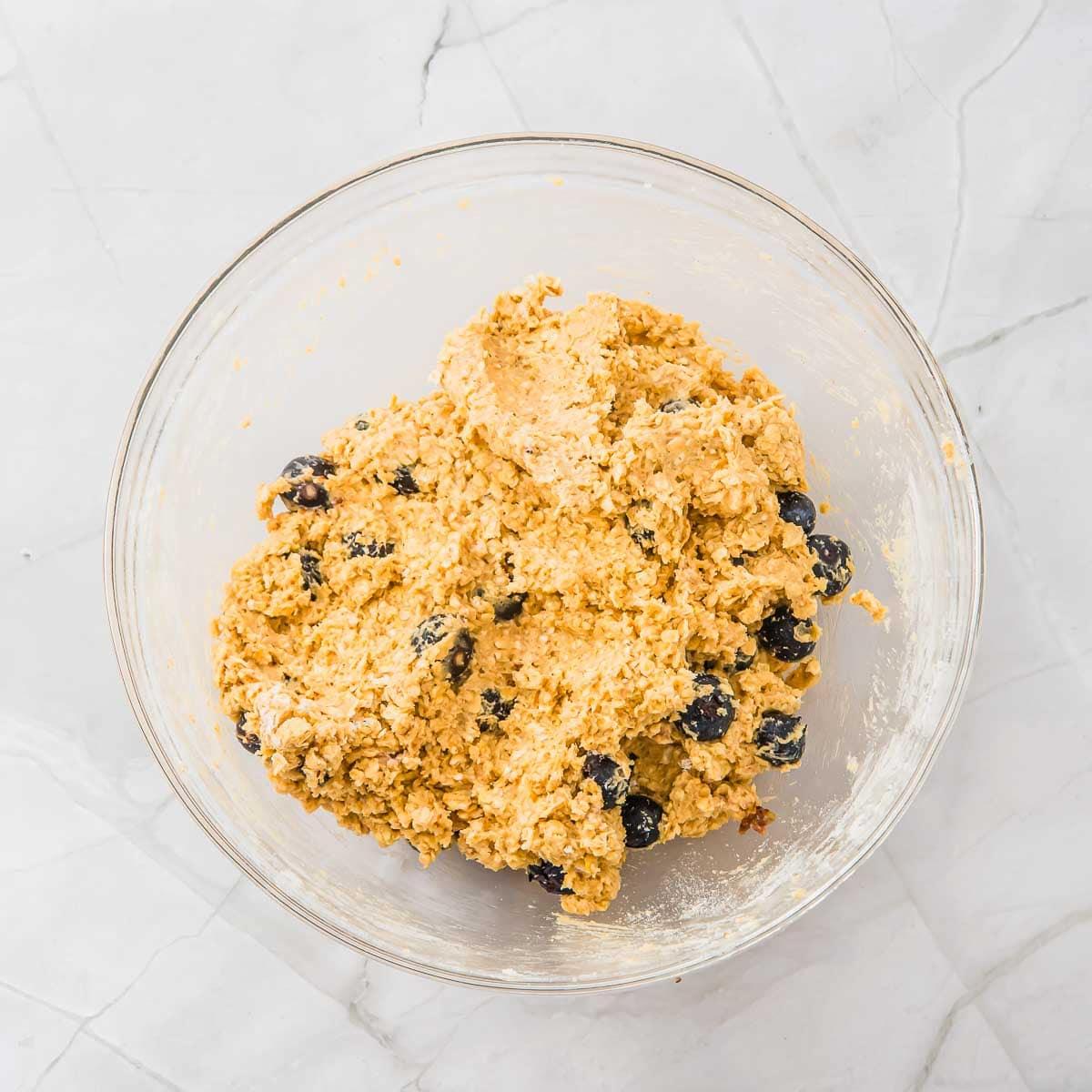 A clear glass bowl on a marble surface filled with thick oatmeal cookie dough mixed with blueberries.