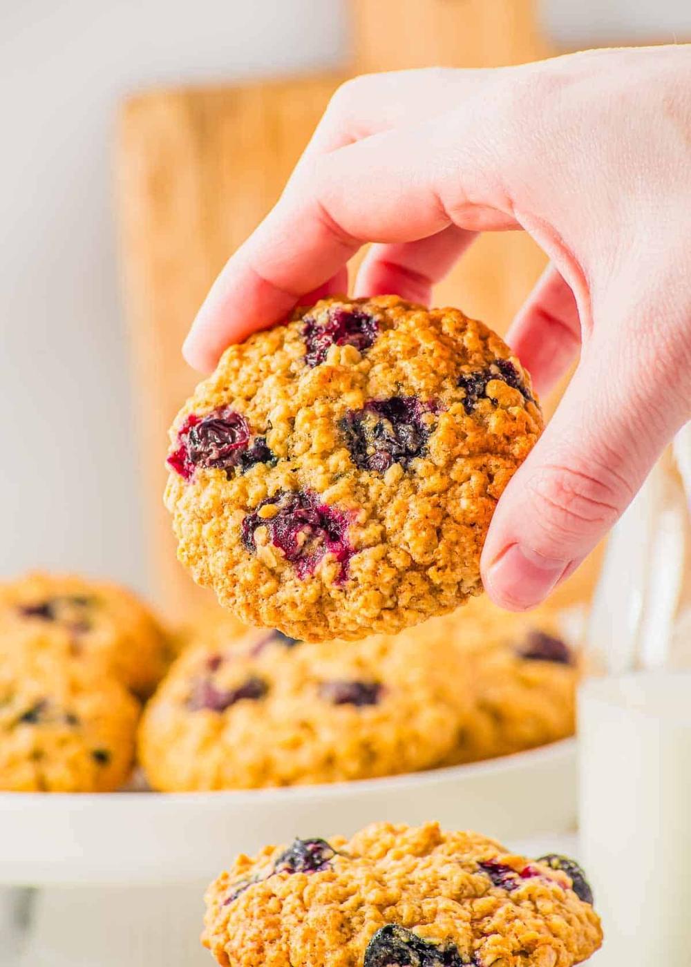 A hand holding an oatmeal cookie with blueberries, with a plate of similar cookies in the background. A glass of milk is partially visible next to the plate.