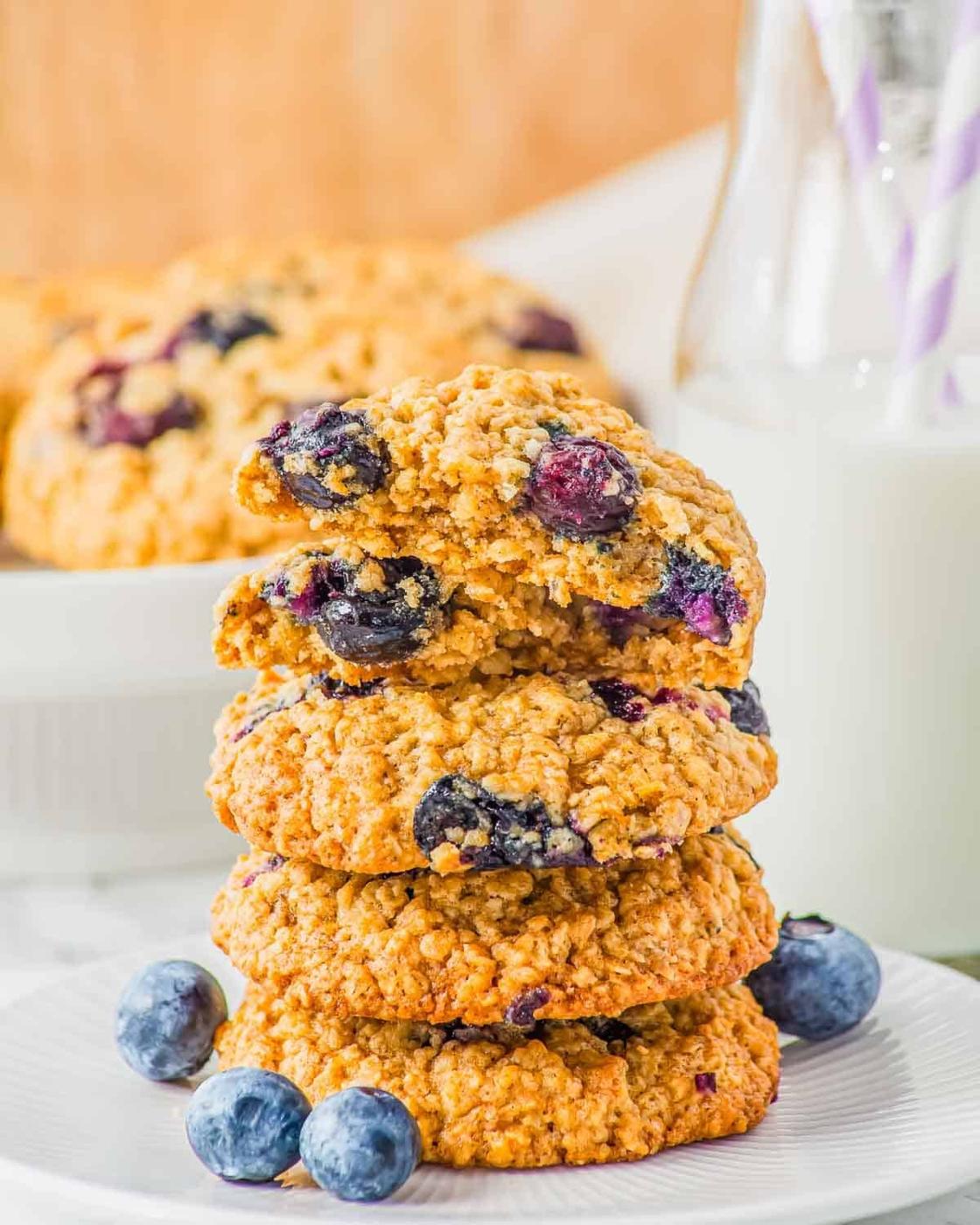 A stack of blueberry oatmeal cookies on a white plate, with fresh blueberries scattered around. In the background, a milk bottle with striped straws is visible, alongside more cookies on a plate.