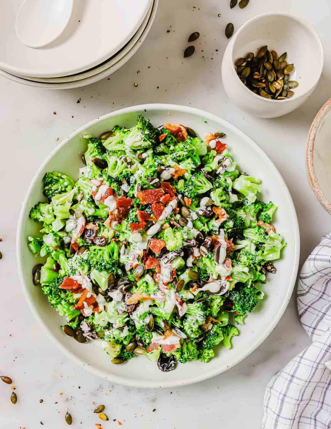 A bowl of broccoli salad with bacon pieces, and raisins sits next to a stack of plates and a small bowl of pumpkin seeds.