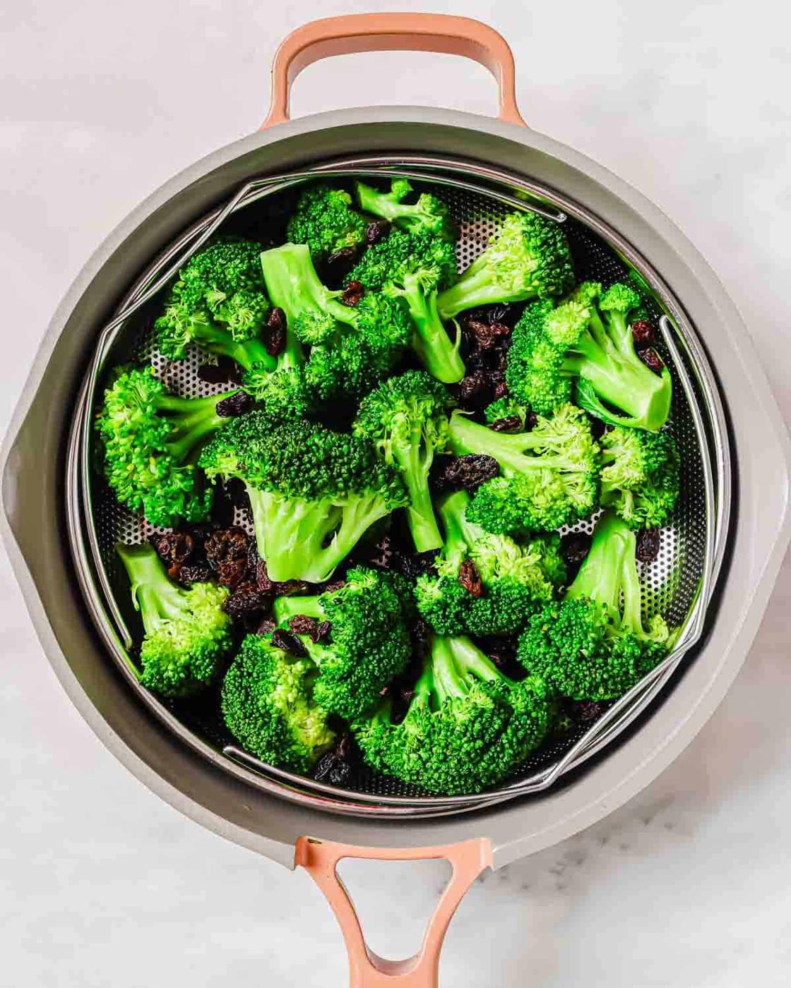Steamed broccoli florets in a metal colander inside a beige pot with copper handles on a light countertop.
