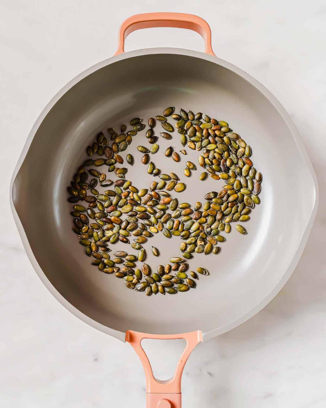 A gray frying pan with a pink handle containing a single layer of roasted pumpkin seeds on a white marble surface.