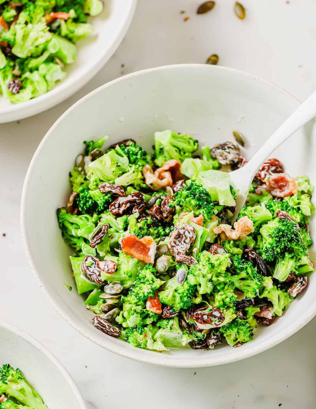 A bowl of broccoli salad with raisins, sunflower seeds, bacon bits, and a creamy dressing, accompanied by a white spoon. Three bowls are partially visible in the background.