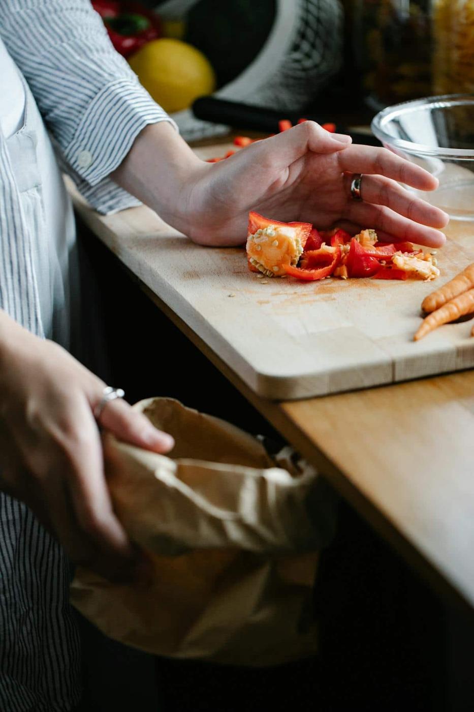 A person preparing food on a wooden kitchen counter, with sliced red peppers and an orange on a cutting board. They are planning to make homemade vegetable broth and are holding a brown paper bag for scraps, wearing a striped shirt. Carrots are also visible nearby.
