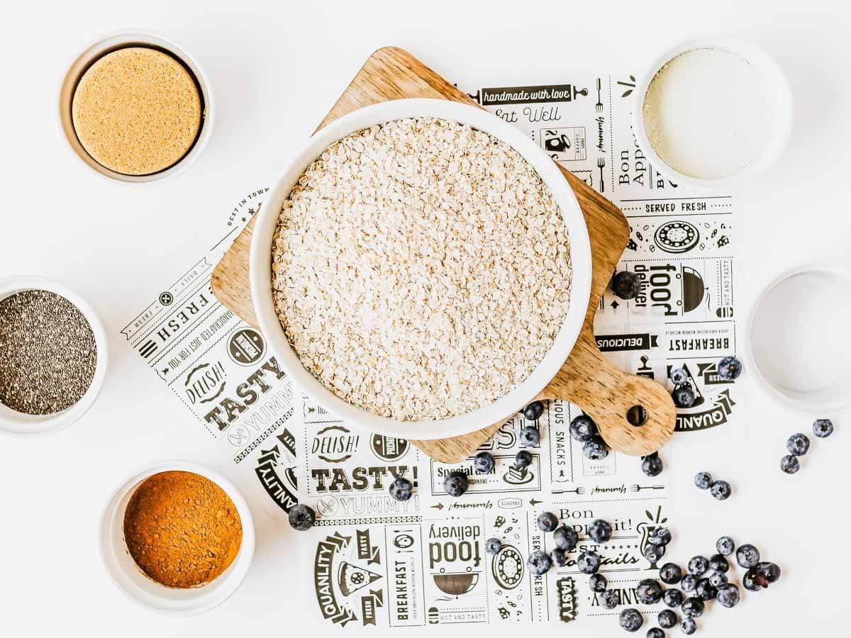 A bowl of dry oats on a wooden board surrounded by bowls of brown sugar, chia seeds, cinnamon, milk, and scattered blueberries, all arranged on a decorative paper background.