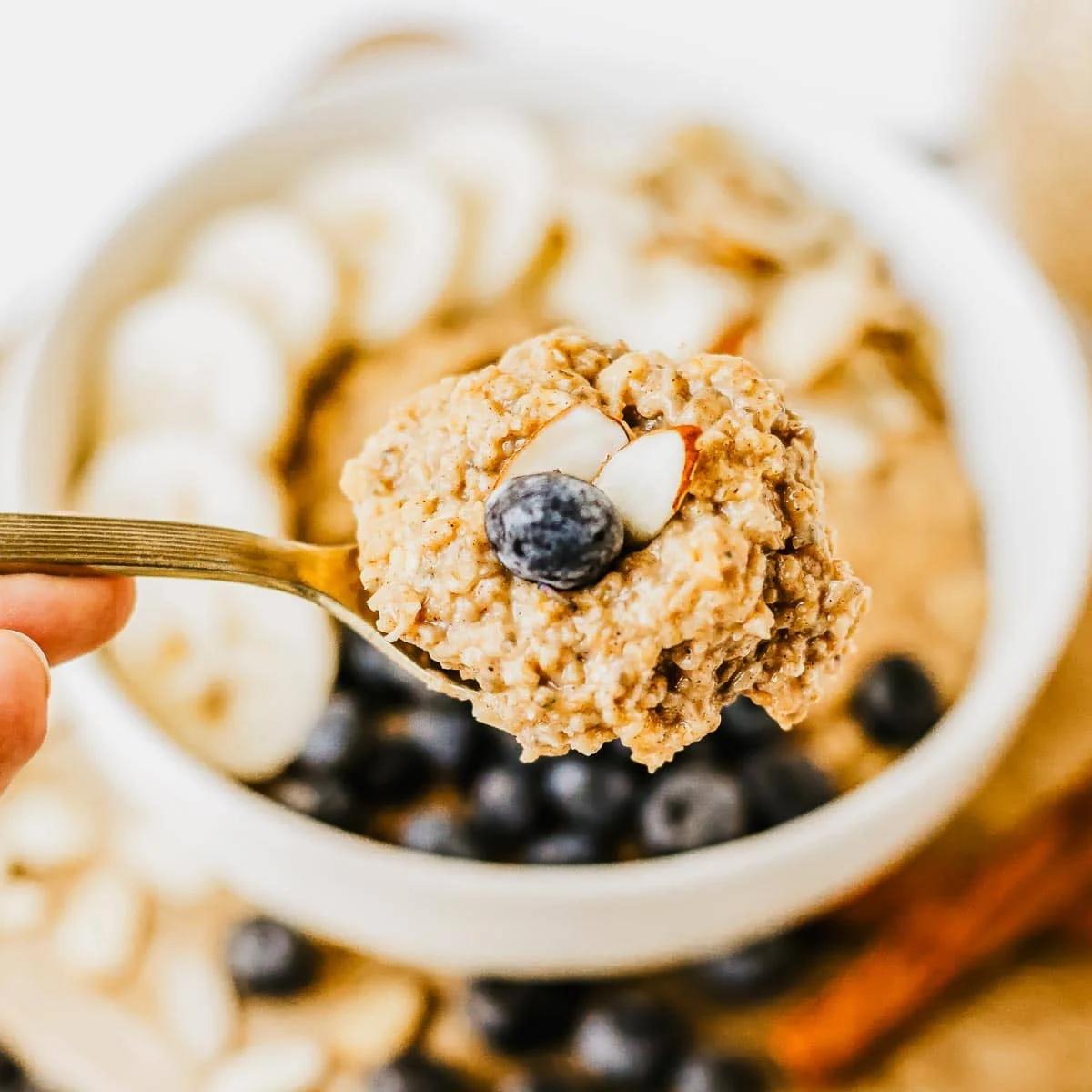 A close-up of a spoonful of creamy brown sugar oatmeal topped with a blueberry and almond slices, held above a bowl of oatmeal garnished with banana slices and more blueberries.