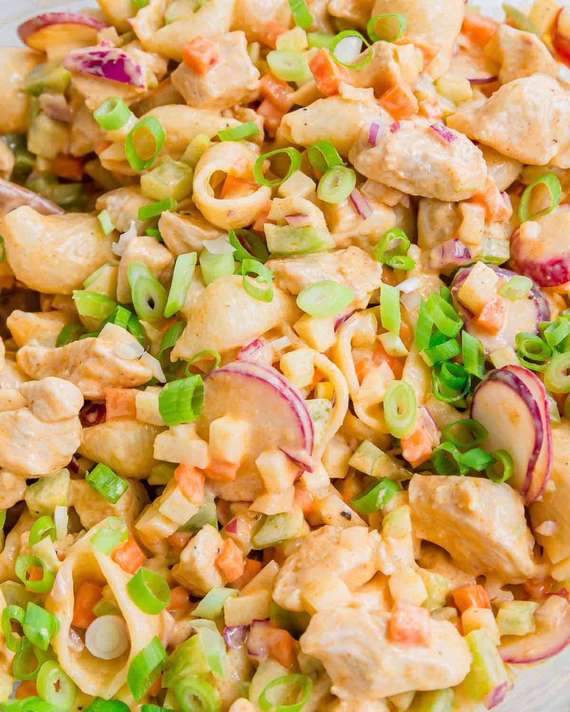 A close-up of a creamy buffalo chicken salad with cooked pasta shells, diced vegetables, radish slices, and chopped green onions sprinkled on top. The salad appears fresh and colorful.