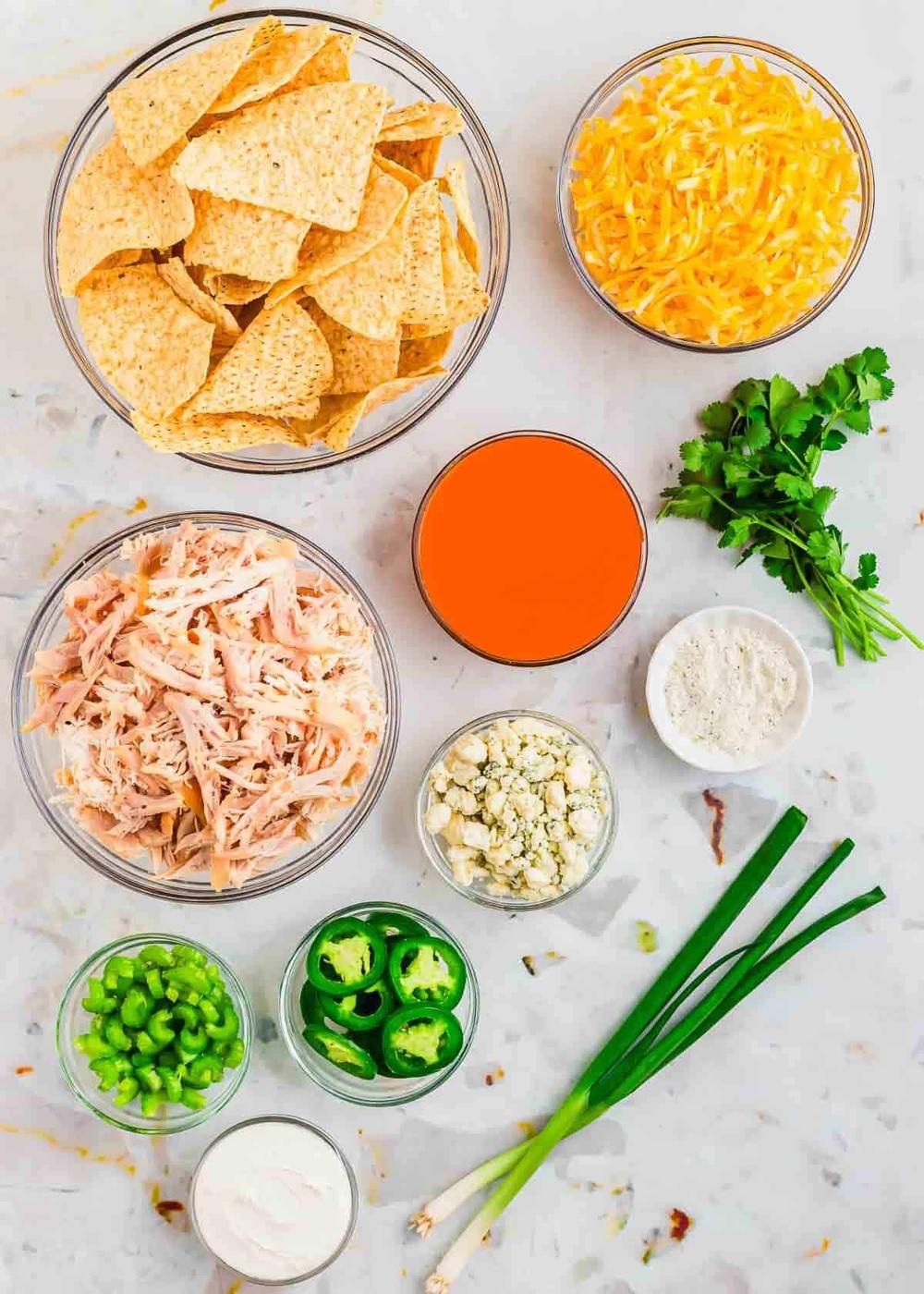 Ingredients for buffalo chicken nachos on a marble surface: tortilla chips, shredded cheddar cheese, cooked shredded chicken, buffalo sauce, fresh cilantro, blue cheese crumbles, sliced jalapeños, green onions, and sour cream.