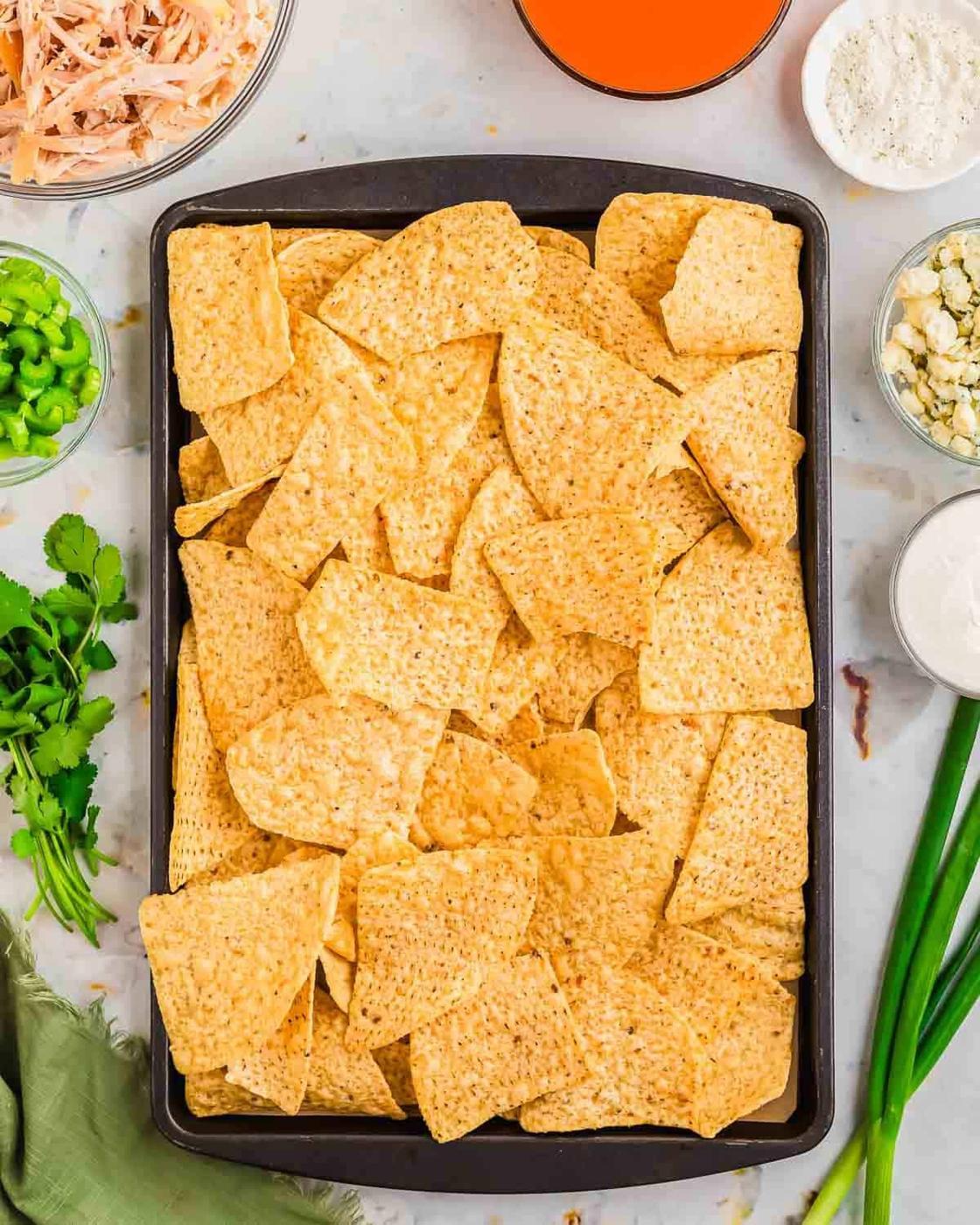 A baking sheet filled with tortilla chips surrounded by bowls of various ingredients including shredded meat, diced green peppers, crumbled cheese, and sauces. Fresh herbs and green onions are placed nearby on a light-colored surface.