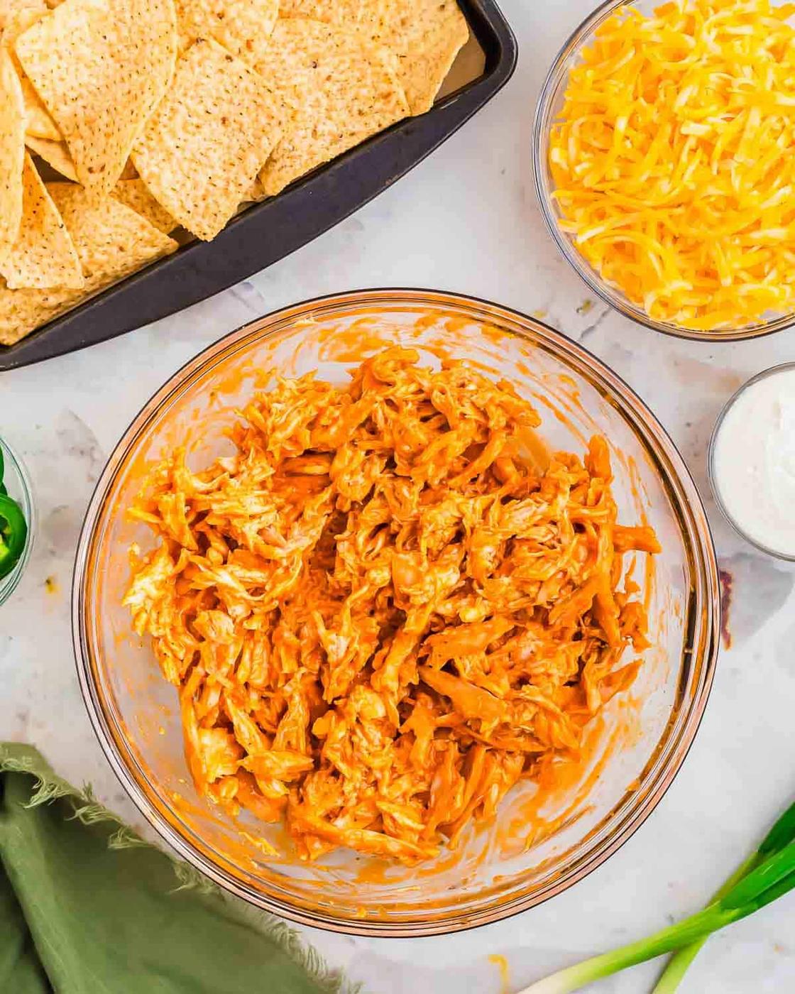A bowl of shredded buffalo chicken sits on a countertop. Surrounding it are dishes with tortilla chips, shredded cheese, and a small bowl of ranch dressing. A green cloth and green onions are visible beside the setup.