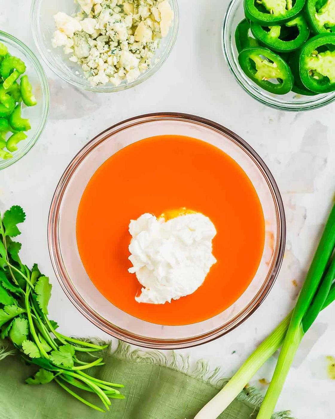 A bowl filled with orange buffalo sauce topped with a dollop of Greek yogurt on a marble surface. Surrounding it are sliced jalapeños, chopped green peppers, blue cheese crumbles, cilantro, and green onions on a green cloth.