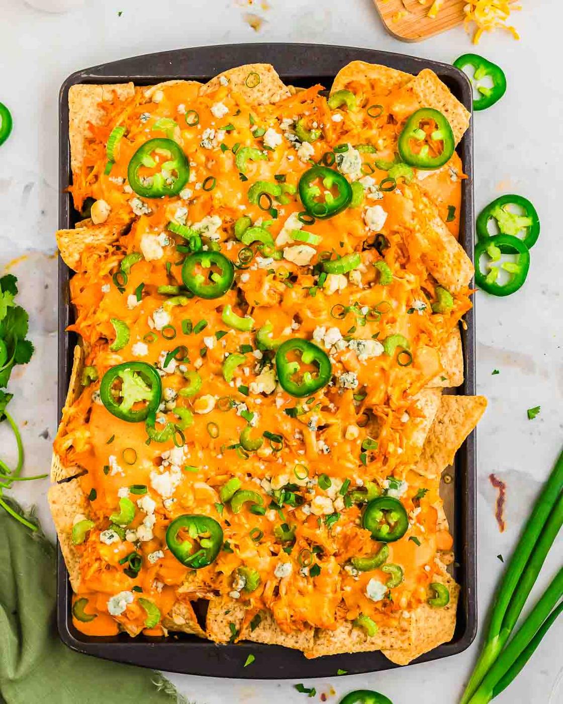A tray of buffalo nachos topped with melted cheese, slices of jalapeño, chopped green onions, and crumbled blue cheese. The tray is placed on a surface with scattered jalapeño slices, cilantro, and a bundle of green onions nearby.