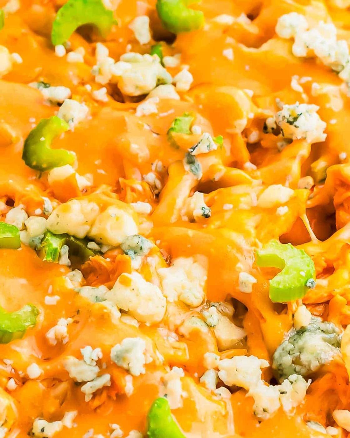Close-up of buffalo chicken nachos, melted cheese, and crumbled blue cheese. Bright orange sauce covers the top, adorned with sliced celery pieces. The textures and colors suggest a flavorful and spicy culinary creation.