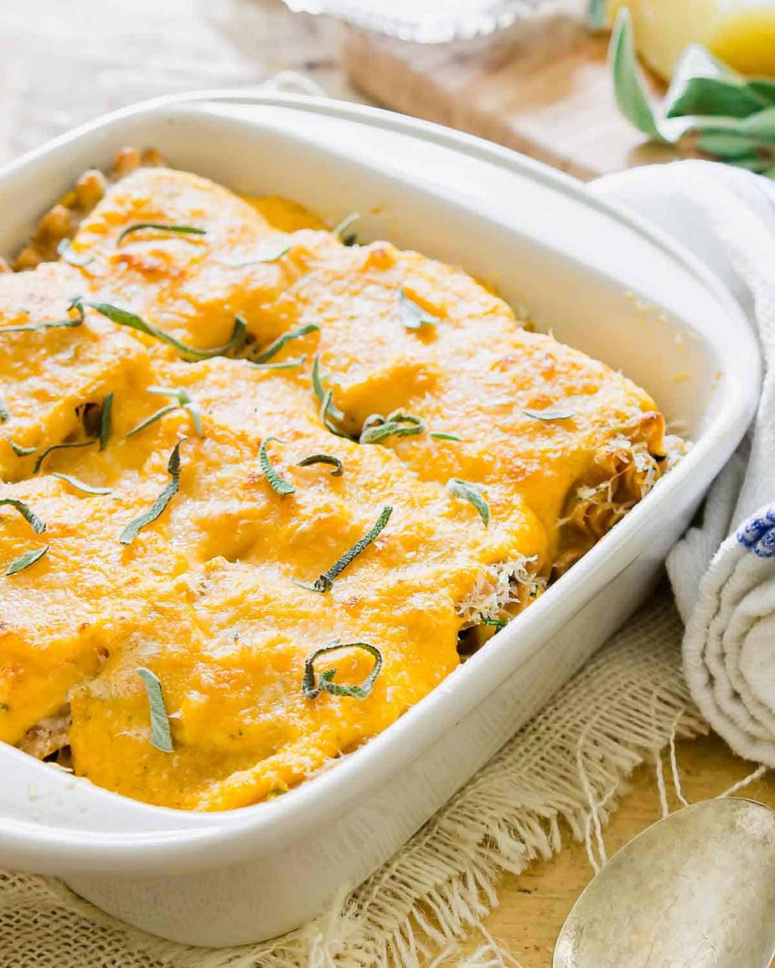 A white rectangular baking dish filled with lasagna rollups topped with a creamy sauce and garnished with sliced fresh herbs. It rests on a rustic cloth next to a folded kitchen towel and spoon.