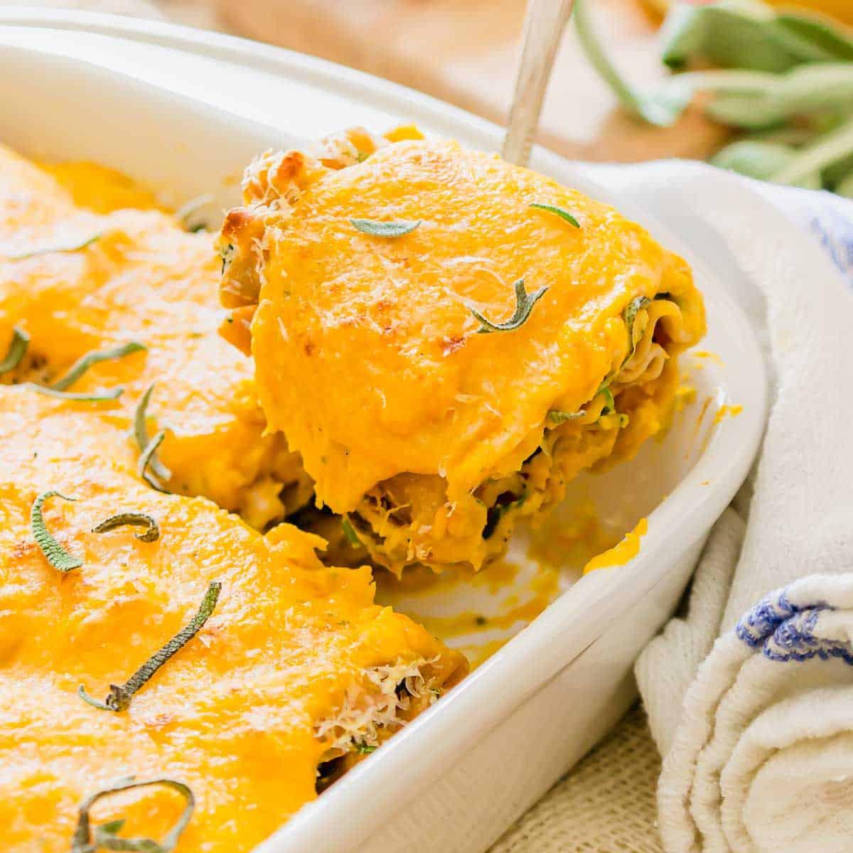 A close-up of a slice of cheesy butternut squash lasagna rollups being lifted from a baking dish. The lasagna is topped with melted cheese and garnished with fresh sage leaves. A white towel is visible in the background.
